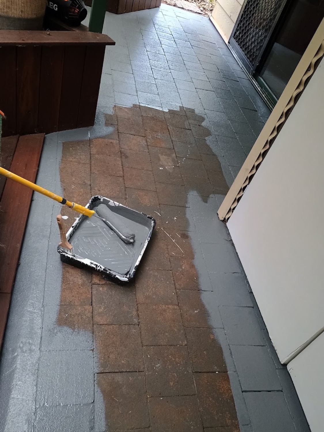 A roller and paint tray on a paved verandah part way through a coat